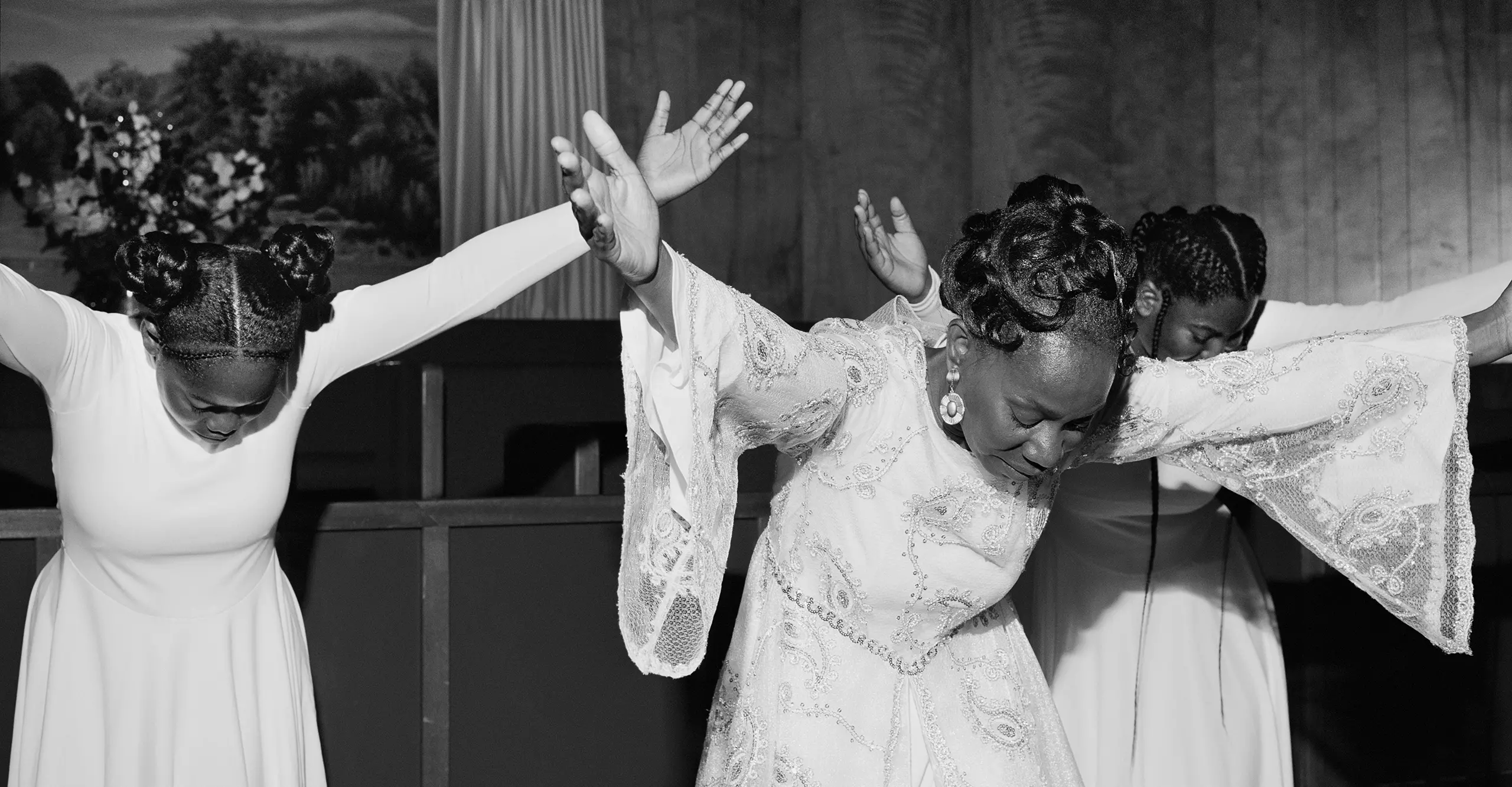 Black and white photograph of three women, wearing white dresses. They have their heads bent down with their arms outstretched. 